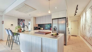 A modern kitchen with a central island, bar stools, a refrigerator, and a backsplash with a mosaic tile design. - Benji Zen Retreat Unique Wavepool Adults Only (Darwin)