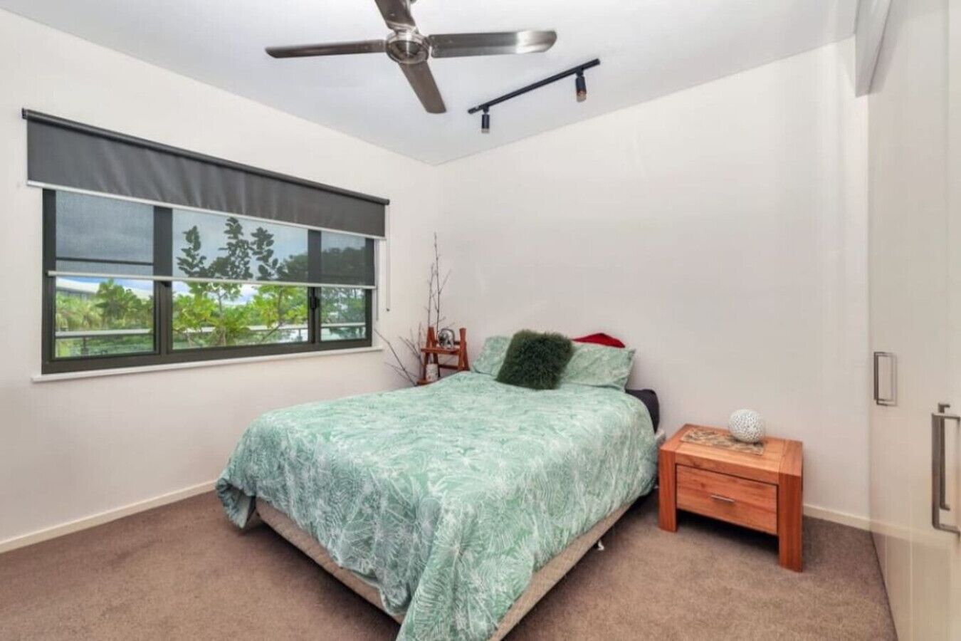 A bedroom with a bed, a nightstand, a ceiling fan, a window with a view of greenery, and a door.