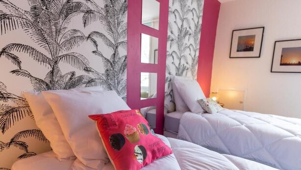 Habitación de hotel con dos camas, ropa de cama blanca, almohada rosa y una pared con estampado de palmeras.