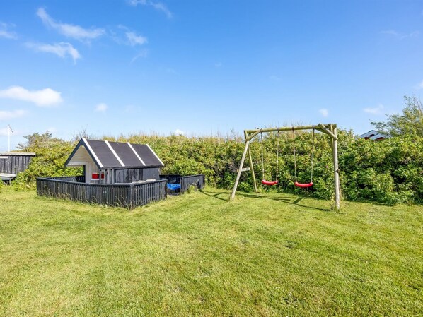 A grassy outdoor area with a wooden swing set, a small shed, and a fenced play area.