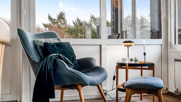A modern room with a large window, a blue armchair, a small table with a lamp, and a candle.