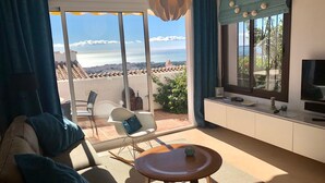 Un soggiorno moderno con un divano, un tavolino e una TV. Si apre una grande finestra con vista sul mare e un balcone.