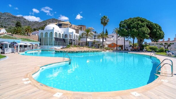 Una grande piscina esterna con un pontile in legno, circondata da edifici con finestre ad arco e palme.