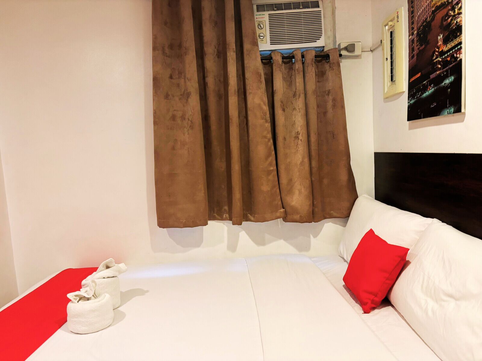 Quarto de hotel com cama, roupa de cama branca, travesseiro vermelho, roupão branco e janela com cortinas castanhas.