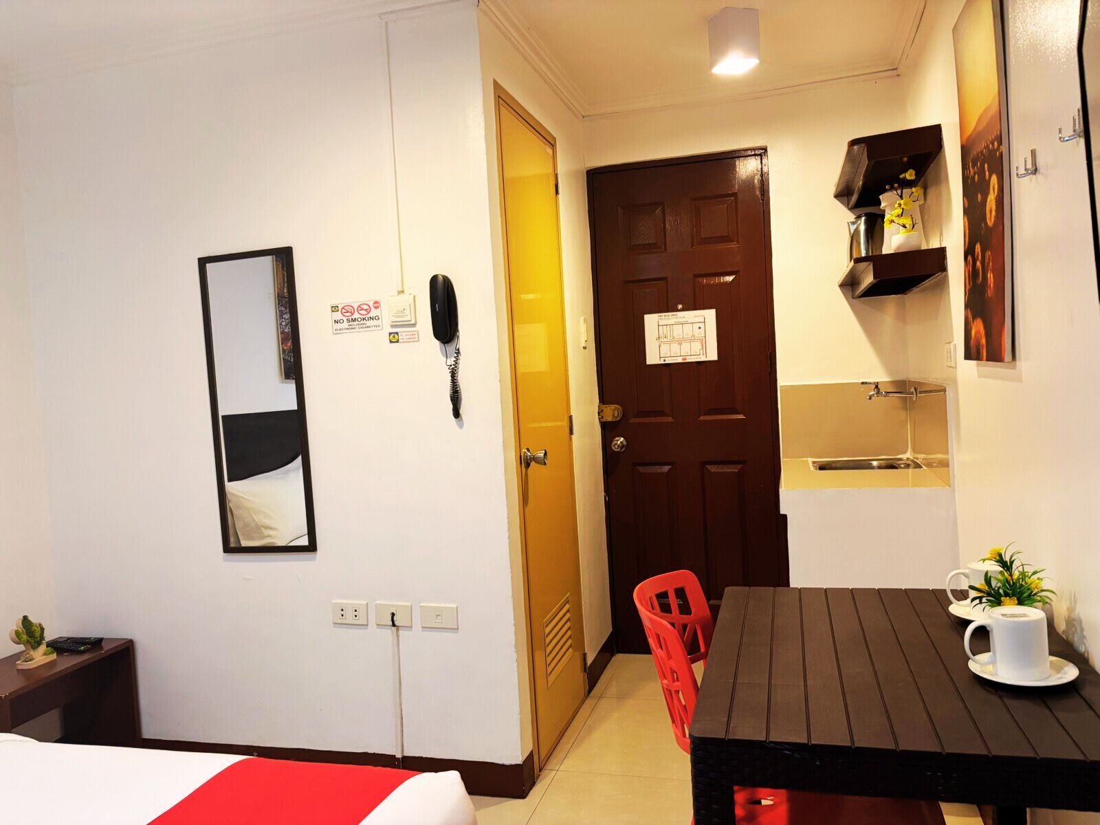 A hotel room with a bed, a desk, a chair, a table, a phone, a mirror, a door, and a small kitchenette.
