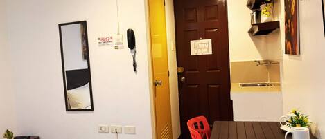 Quarto de hotel com cama, escrivaninha, cadeira, mesa, telefone, espelho, porta e pequeno espaço de cozinha.