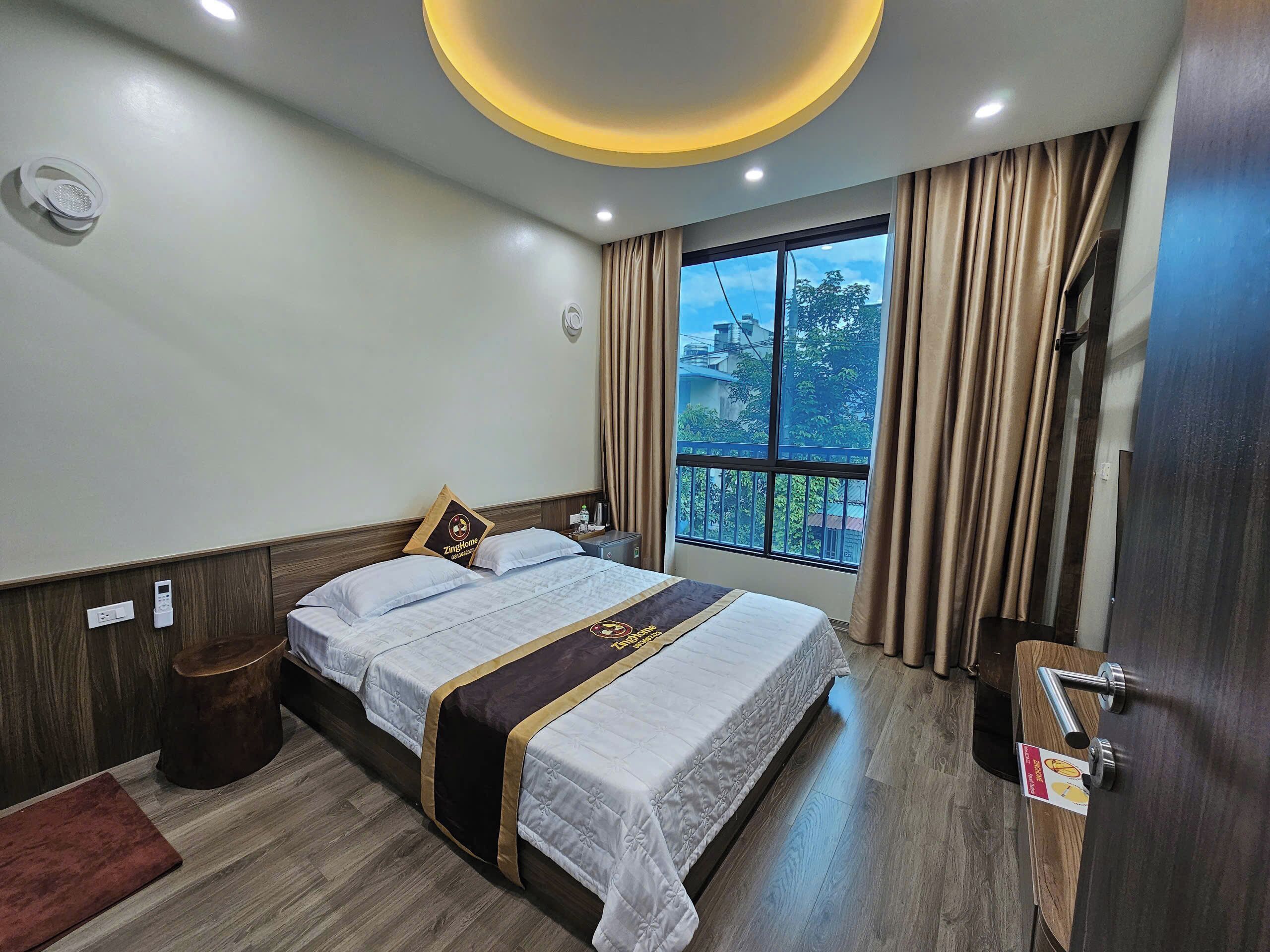A hotel room with a bed, a bedside table, a nightstand, a window with curtains, and a ceiling light fixture.
