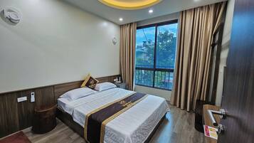 A hotel room with a bed, a bedside table, a nightstand, a window with curtains, and a ceiling light fixture.