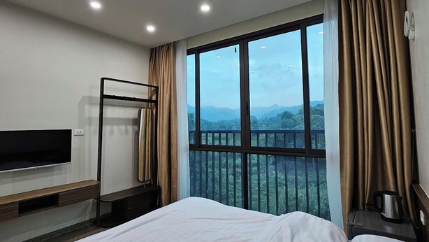 A hotel room with a large bed, a TV on a stand, a balcony with a view of the mountains, and a coffee maker on a nightstand. - ZingHome Ha Giang (Ha Giang 1)