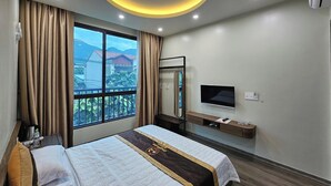 A hotel room with a large bed, a TV, a mirror, and a window with a view of buildings and mountains. - ZingHome Ha Giang (Ha Giang 1)