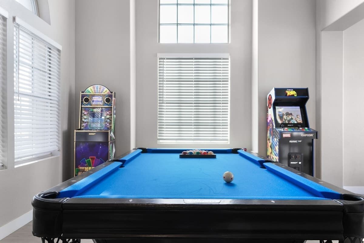 A pool table with a blue felt surface, surrounded by windows and arcade machines.