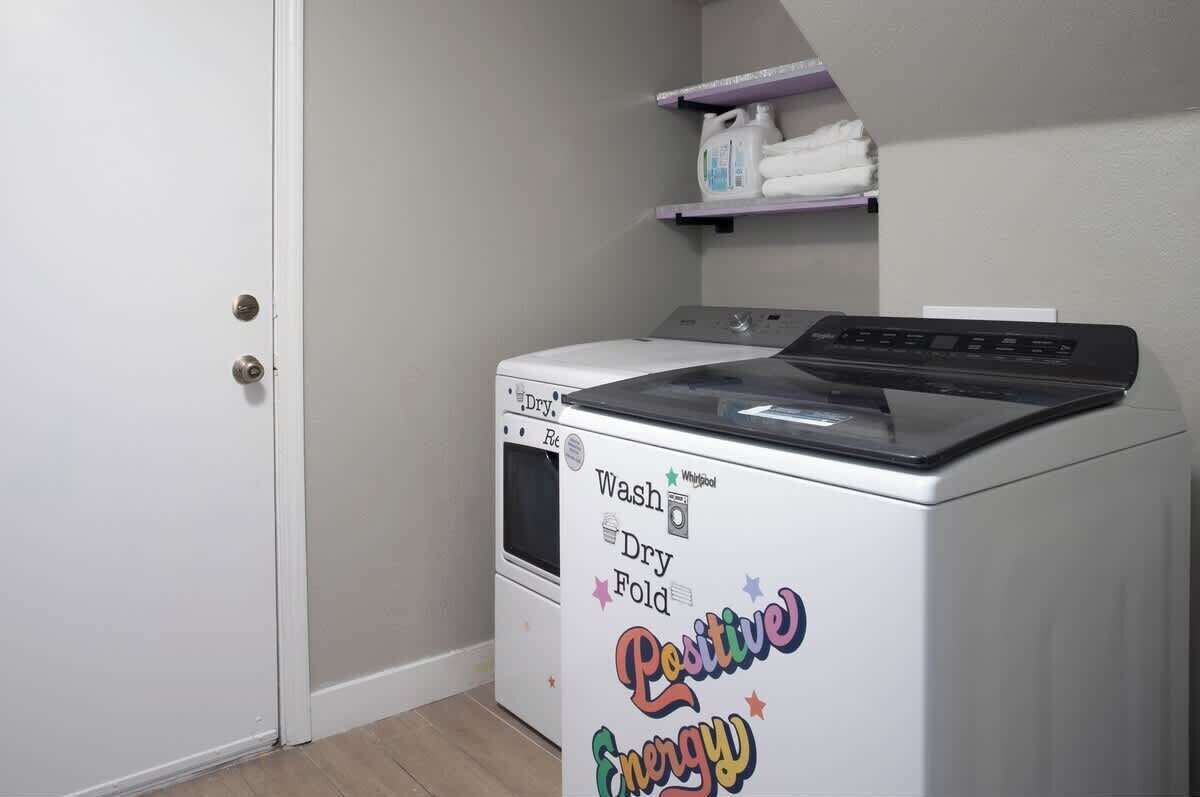Une petite machine à laver à chargement frontal, de couleur blanche, dotée d’un panneau de commande. Elle se trouve dans une petite buanderie avec une porte blanche et une étagère au-dessus, où sont pliés les serviettes.