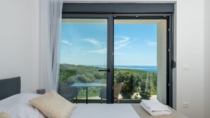 A bedroom with a bed, a table, and chairs, a balcony with a table and chairs, and a view of greenery and water. - Villa Artec with Sea View and Private Pool (Marcana)