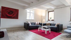 A modern living room with a dark grey sofa, a glass coffee table, a red rug, and a grey armchair. There are two windows with white frames and a red rose painting on the wall.