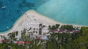 Vista aérea de un complejo turístico en la playa, con una playa de arena, palmeras y un estacionamiento con carpas blancas.