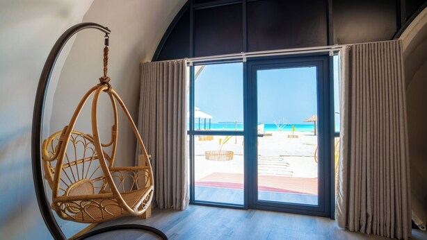 Una habitación con una silla de mimbre, un ventanal con cortinas y vista a la playa.