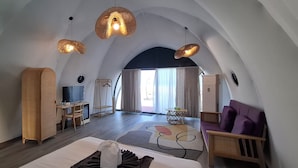 Una habitación de hotel moderna con techo abovedado, cama con cabecera blanca y marrón, zona de estar con sofá púrpura y mesa de comedor con sillas.