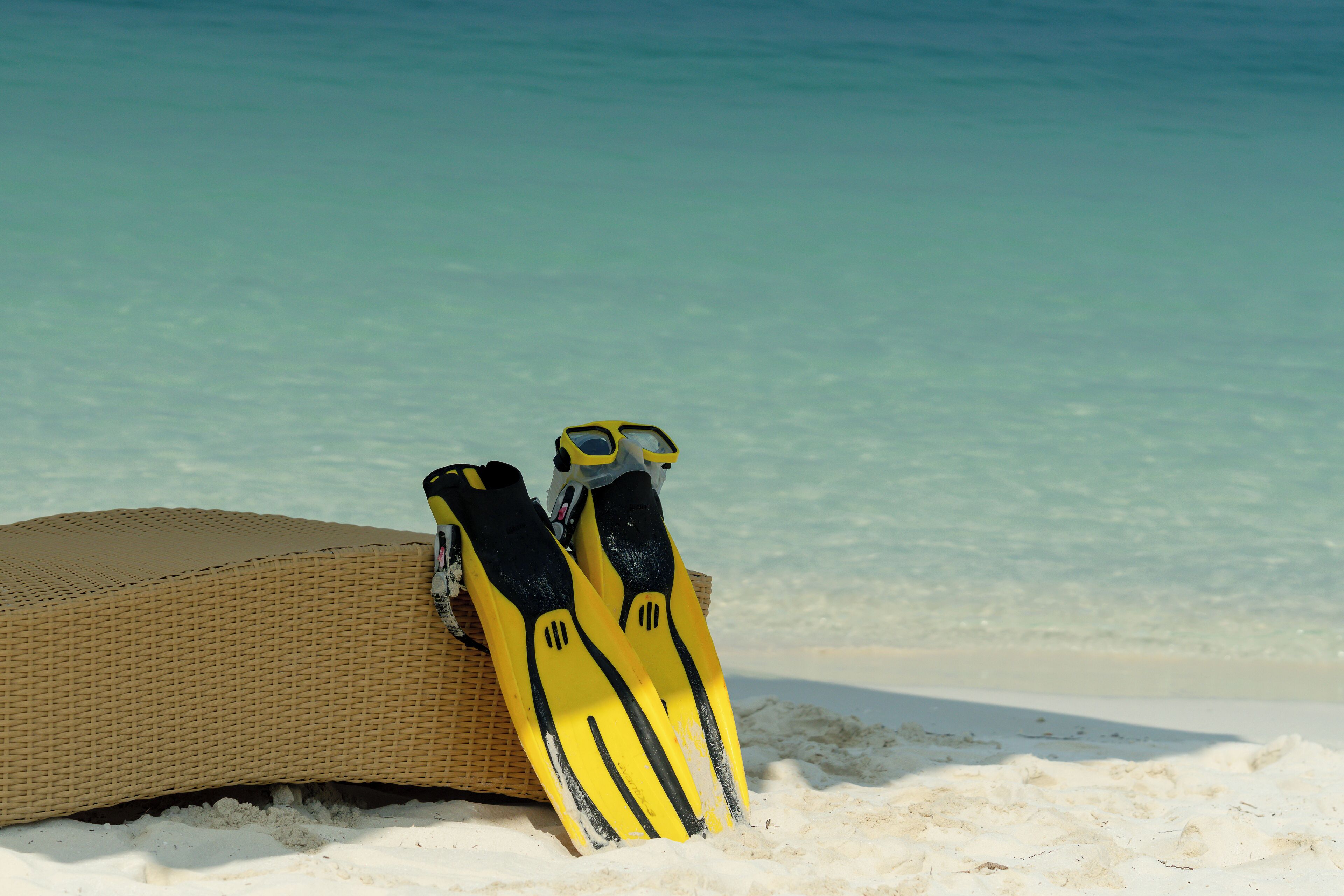 A beach scene with a wicker lounge chair and yellow flippers leaning against it.