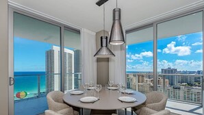 Un coin repas avec une table ronde dressée pour six personnes, de grandes fenêtres offrant une vue sur la ville et deux lampes suspendues.