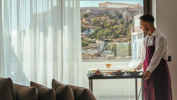 A modern room with a large window overlooking a historic cityscape and a table with a glass of wine and a plate of food. - Eternity Athens (Athens)