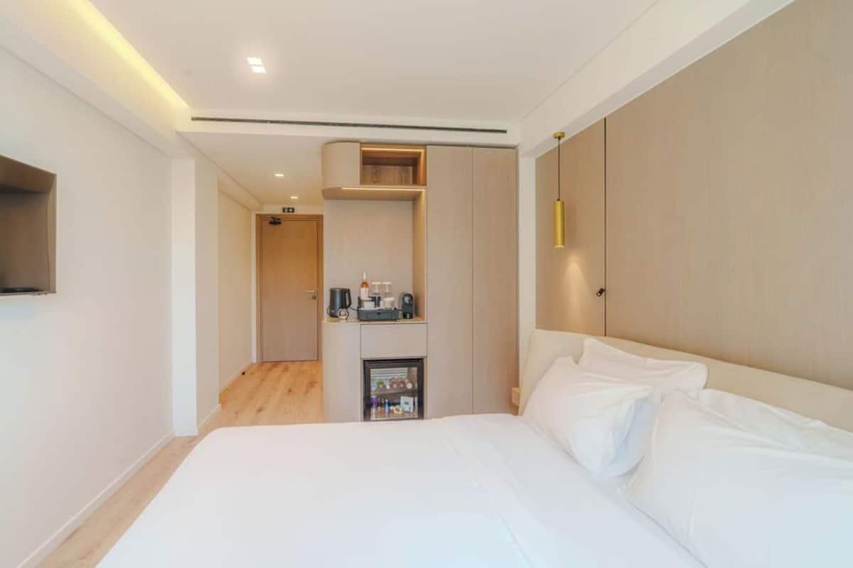 A modern hotel room with a large bed, a built-in wardrobe, a minibar, and a flat-screen TV mounted on the wall.