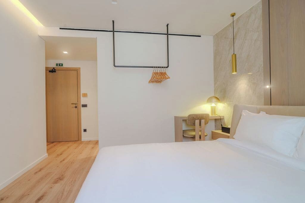 Et moderne hotelværelse med trægulv, en seng med hvide sengetæger, et natbord med en lampe og et håndklædestativ på væggen.