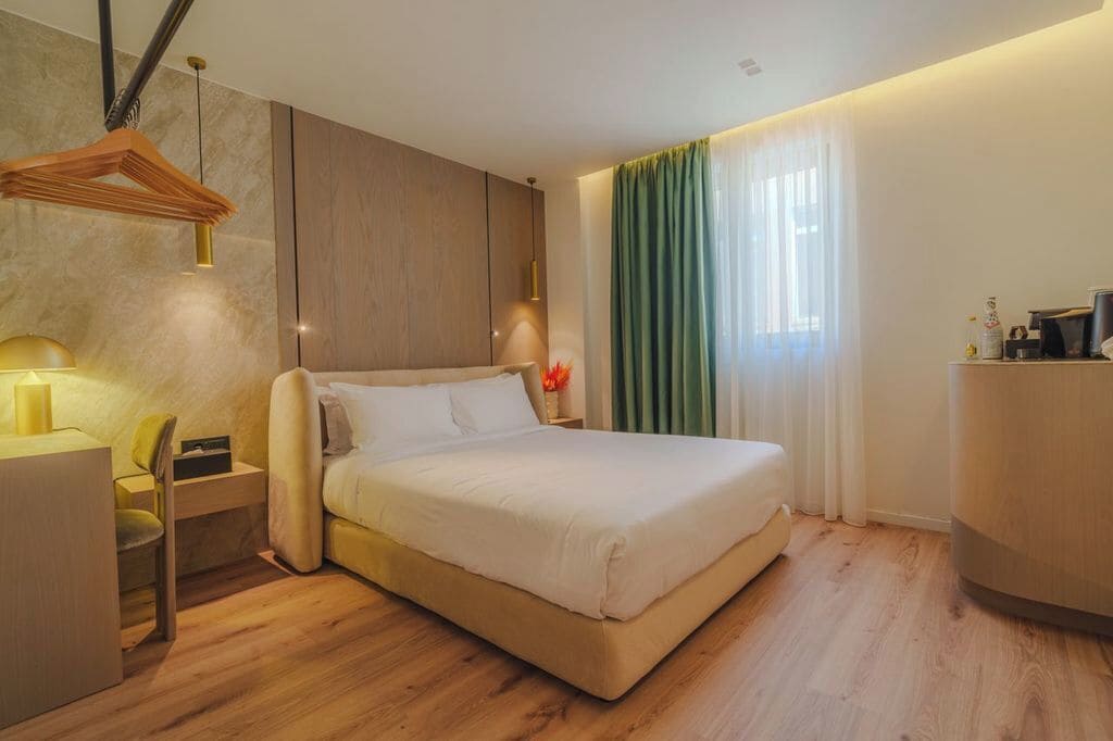 Quarto de hotel moderno com cama, mesa de cabeceira, abajur e escrivaninha com cadeira.