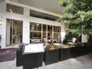 A modern hotel exterior with a glass facade, outdoor seating area, and a signboard.