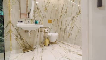 A bathroom with a marble floor and walls, a white sink, a toilet, and a gold-colored trash bin.