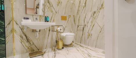 A bathroom with a marble floor and walls, a white sink, a toilet, and a gold-colored trash bin.