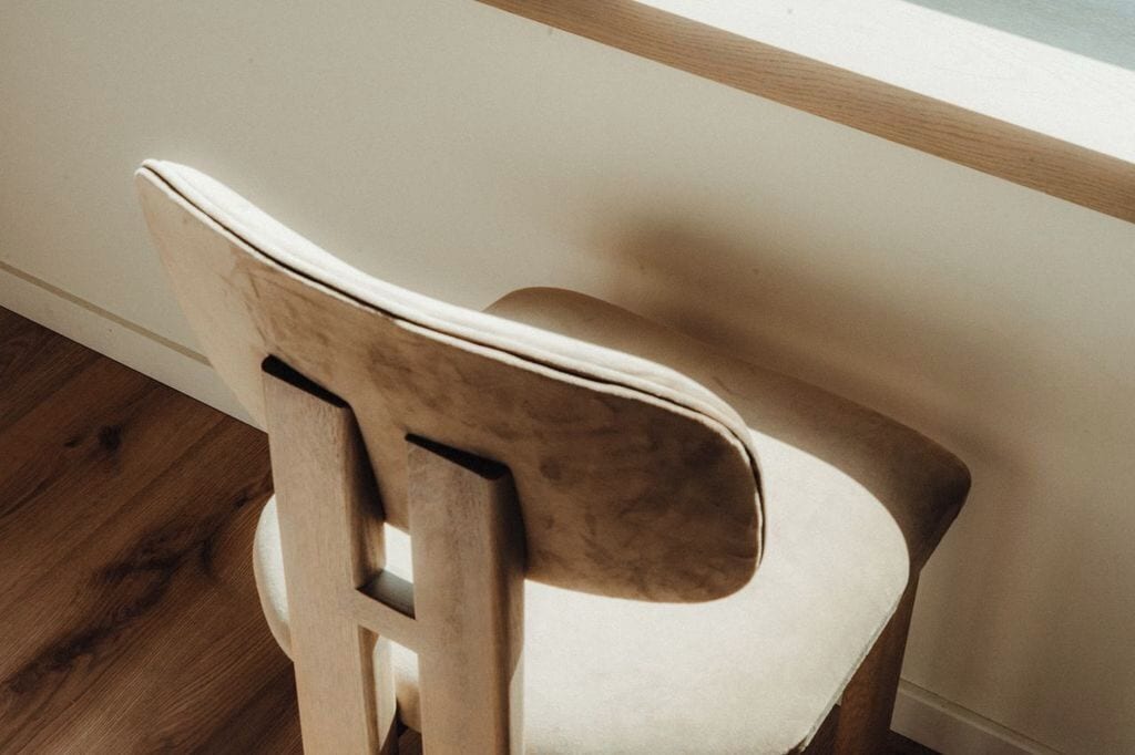 Una silla de madera con asiento y respaldo acolchados, ubicada frente a una pared con un ventanal.