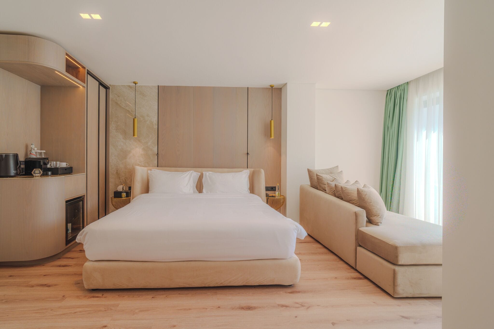 A modern hotel room with a large bed, a sofa, a small table with a coffee maker, and a window with curtains.