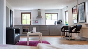 Una cocina moderna con armarios grises, encimera blanca y horno empotrado. En la sala de estar, un sofá de cuero negro y una mesa blanca con un tapete púrpura.