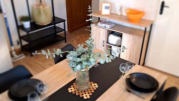 Un comedor con una mesa de madera dispuesta para cuatro, un florero con follaje y un mantel individual negro.