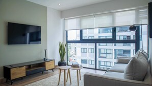 Ein modernes Wohnzimmer mit Flachbildfernseher, einer Couch, einem Couchtisch und einem großen Fenster mit Blick auf die Stadt.