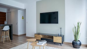 Ein modernes Wohnzimmer mit einem Flachbildfernseher an der Wand, einer Holz-Unterhaltungseinrichtung, zwei Hocker, einer Topfpflanze und einer sichtbaren Tür mit einem Warnschild.