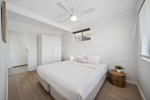 A bedroom with a bed, a ceiling fan, a window, and a small wooden side table.