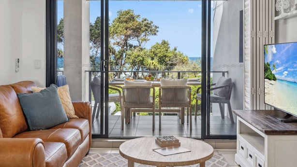 Un salon avec un canapé en cuir, une table basse ronde, un téléviseur et un balcon offrant une vue sur la mer et les arbres.