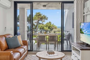 Een woonkamer met een leren bank, een ronde salontafel, een televisie en een balkon met uitzicht op zee en bomen.