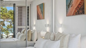 Une chambre à coucher avec un lit, des oreillers, une table de chevet, un miroir et une vue sur l’océan.