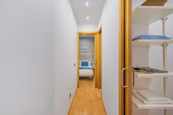 A narrow hallway with wooden flooring leading to a bedroom with a bed, a nightstand, and a shelf with a towel and shoes.