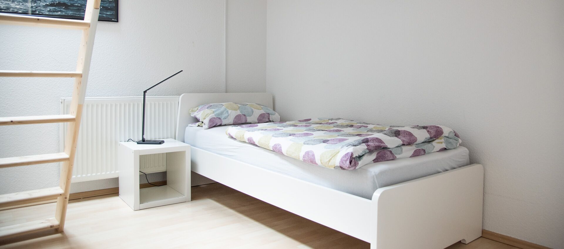 Una habitación pequeña con cama individual, paredes blancas, una escalera apoyada en la pared, una mesita de noche y un radiador.