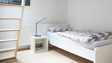 Una habitación pequeña con cama individual, paredes blancas, una escalera apoyada en la pared, una mesita de noche y un radiador.
