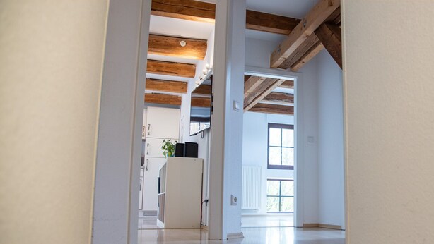 Een gang met zichtbare houten balken, witte muren en een deur die leidt naar een kamer met een radiator en een raam.