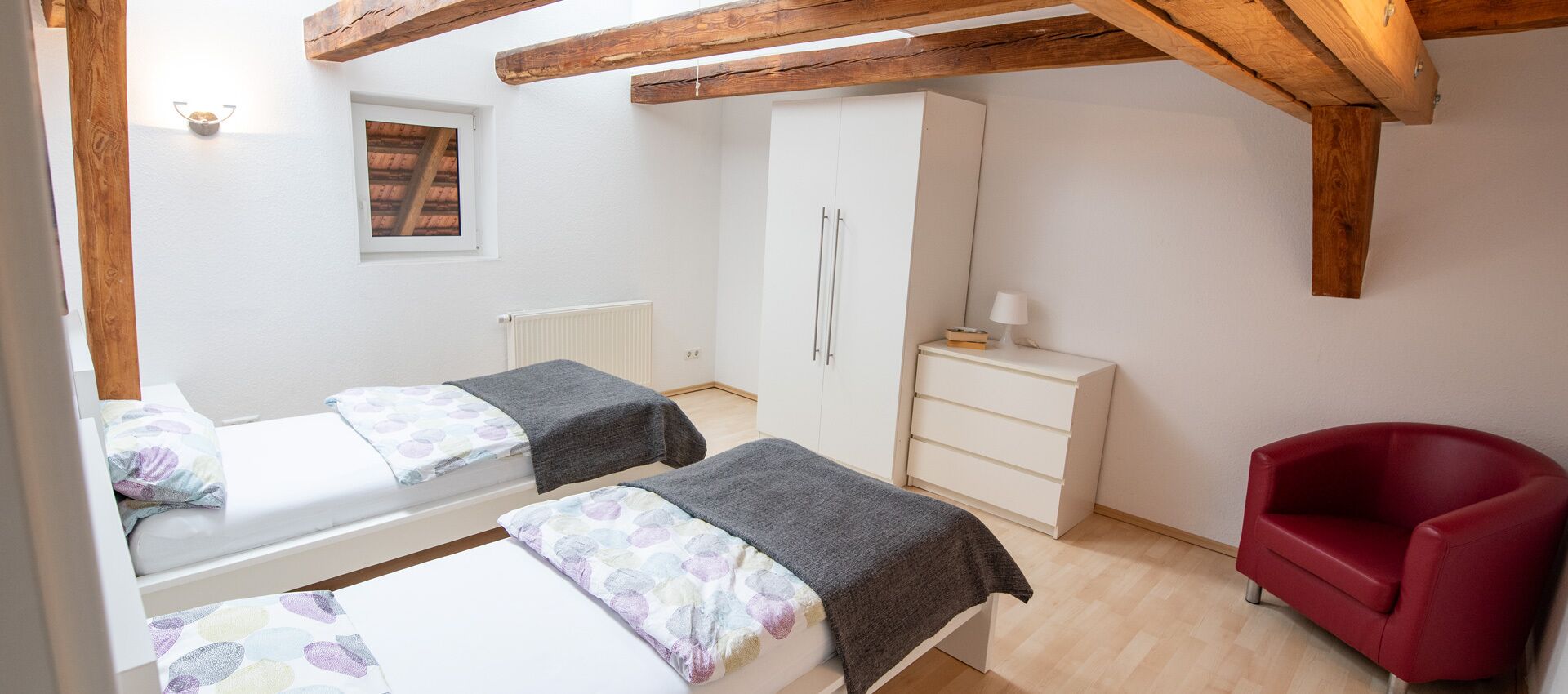 A bedroom with two beds, a wardrobe, a red chair, and a radiator.