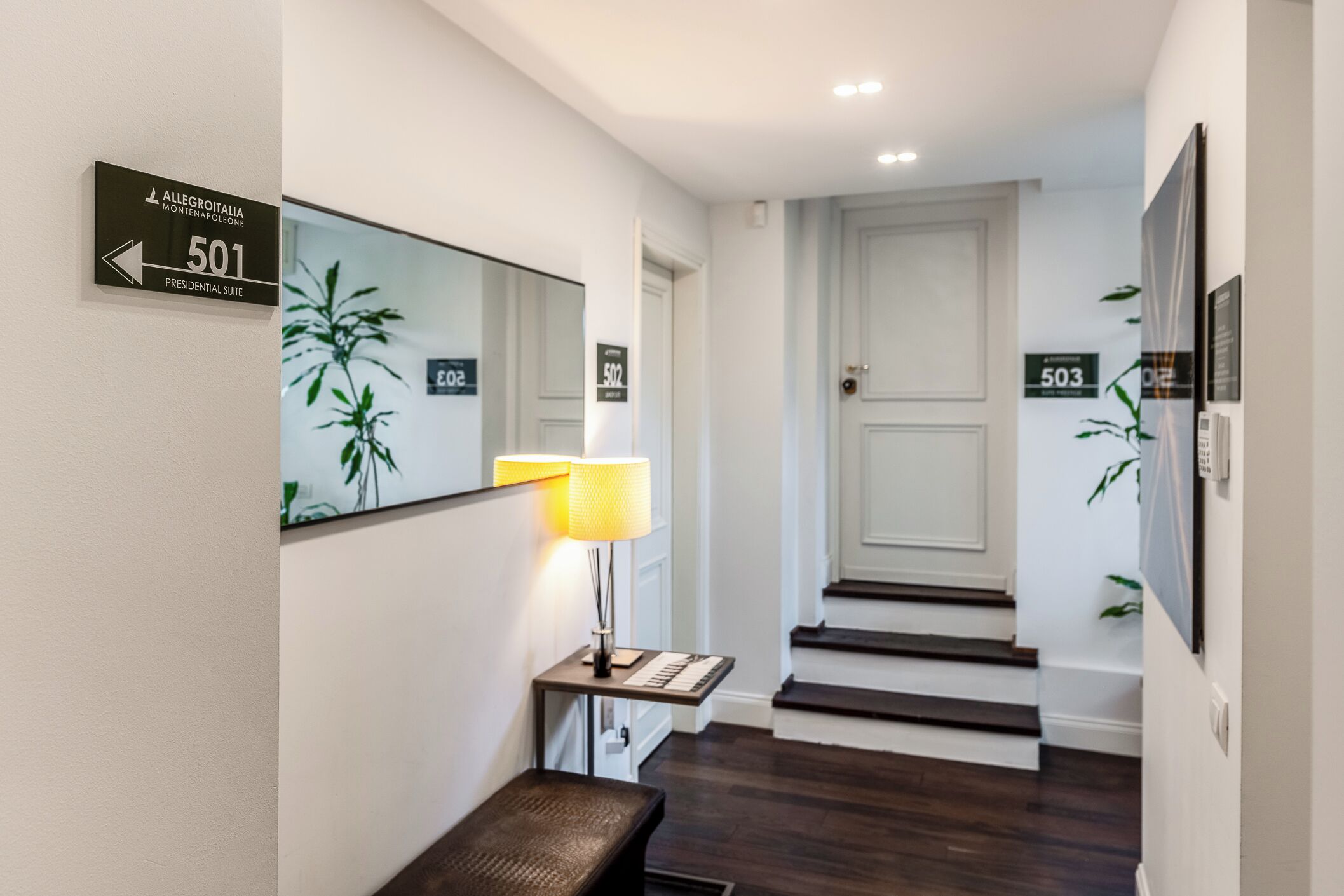 Un couloir d’hôtel avec une pancarte indiquant le numéro de chambre 501, une lampe, un banc et un escalier menant à une autre chambre.
