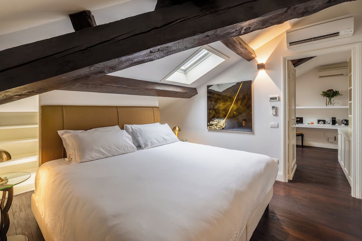 A bedroom with a large bed, a skylight, a bedside table, and a view of a kitchen area.