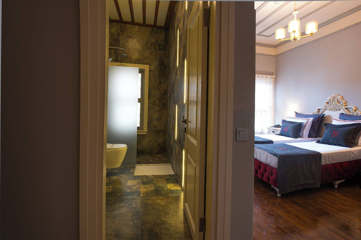 A hotel room with a wooden floor, a bed with a red headboard, and a bathroom with a toilet and shower.