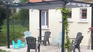 Un área de estar al aire libre con sillas y una mesa bajo una pérgola, rodeada por un camino de grava y follaje.