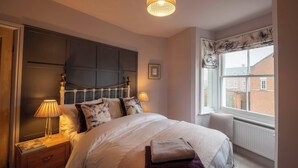 A bedroom with a large bed, a nightstand with a lamp, a window with a view of buildings, and a ceiling light fixture. - Refurbished Victorian House (Colchester)